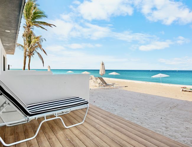 A beach view with a modern white lounger on a deck, palm trees, and a vast ocean under a blue sky with scattered clouds.